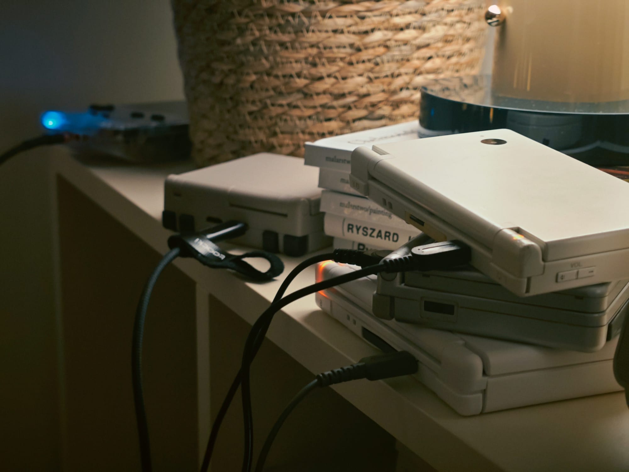 A collection of gaming devices, including a stack of Nintendo DS consoles and a small retro console, are charging on a shelf
