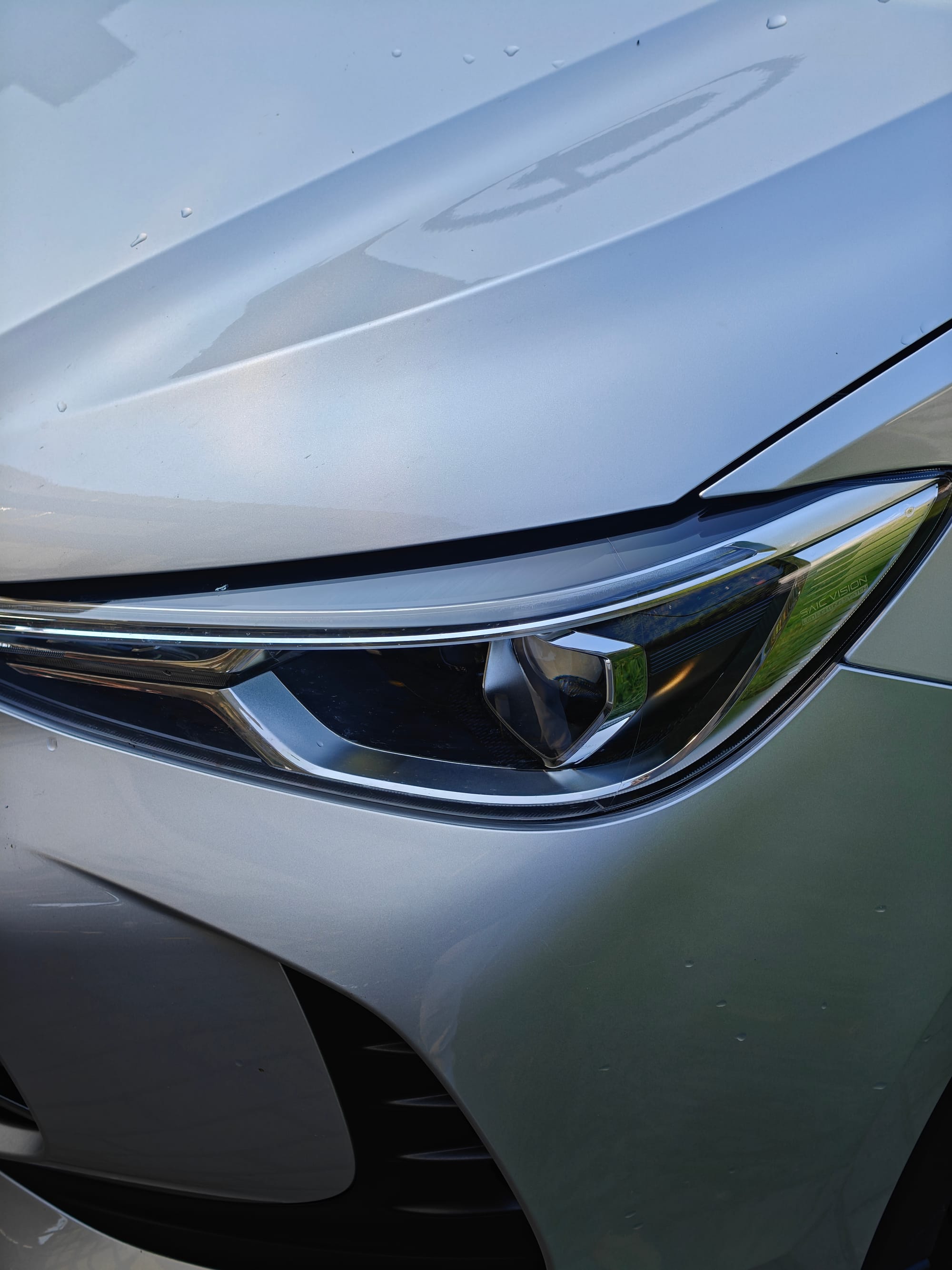 A silver car's front right headlight and part of its hood and bumper are visible, with water droplets on the surface